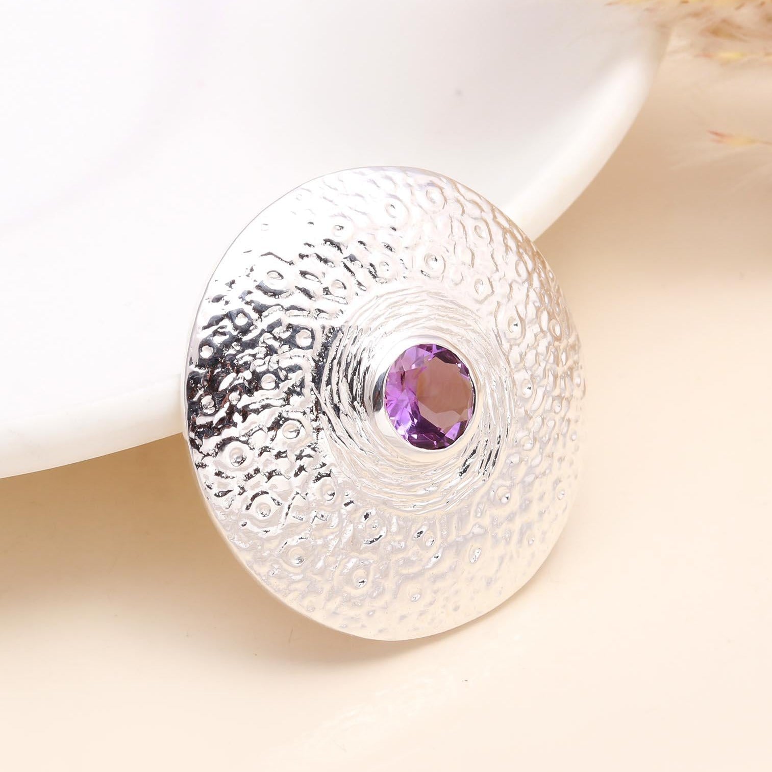 A 925 Sterling Silver Amethyst Textured Disc Pendant placed on a white surface, with the pendant displaying a hand-textured pattern and a natural amethyst gemstone in the center.