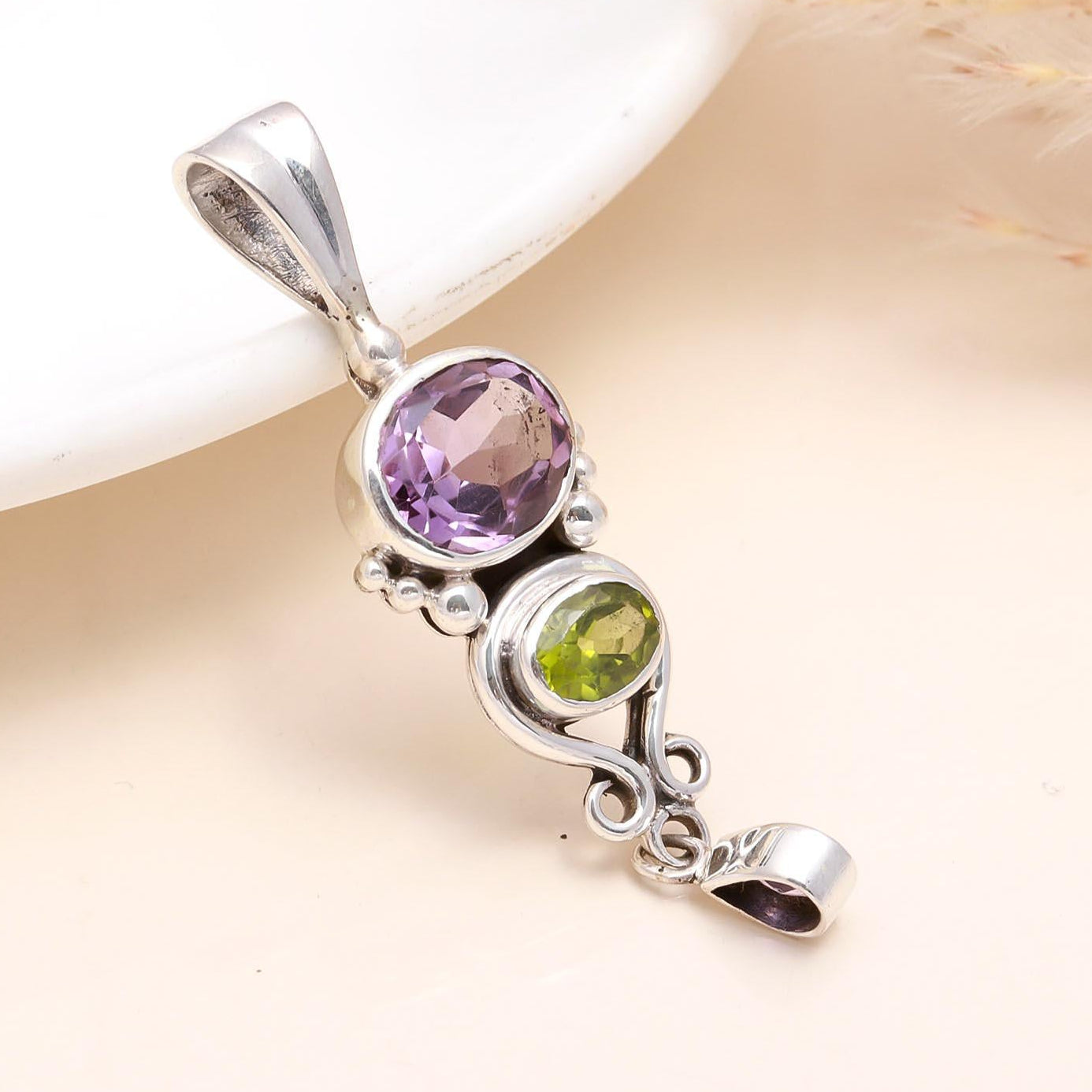 A sterling silver pendant featuring a round amethyst gemstone above a peridot gemstone, set against a neutral background.