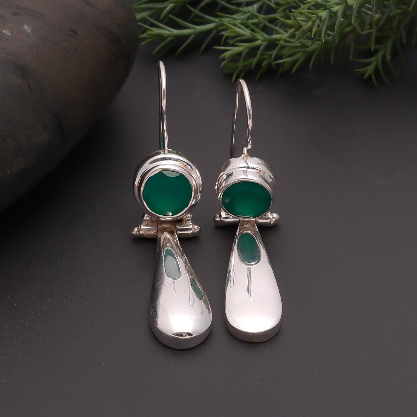 Silver earrings with green stones on a dark surface with greenery in the background