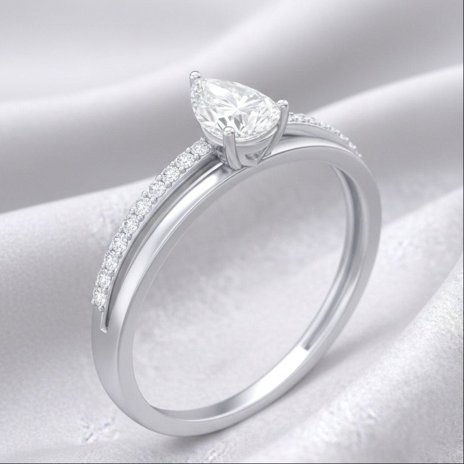 A silver-toned solitaire ring featuring a pear-cut clear gemstone in a prong setting, with a pavé band accented by small round stones, displayed on a draped white satin fabric background.