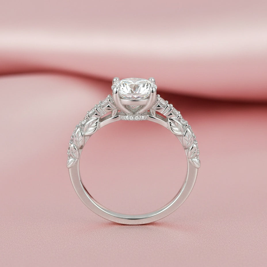 A silver-toned solitaire engagement ring with a round-cut center stone and decorative pavé-set side stones, displayed on a soft pink satin background.