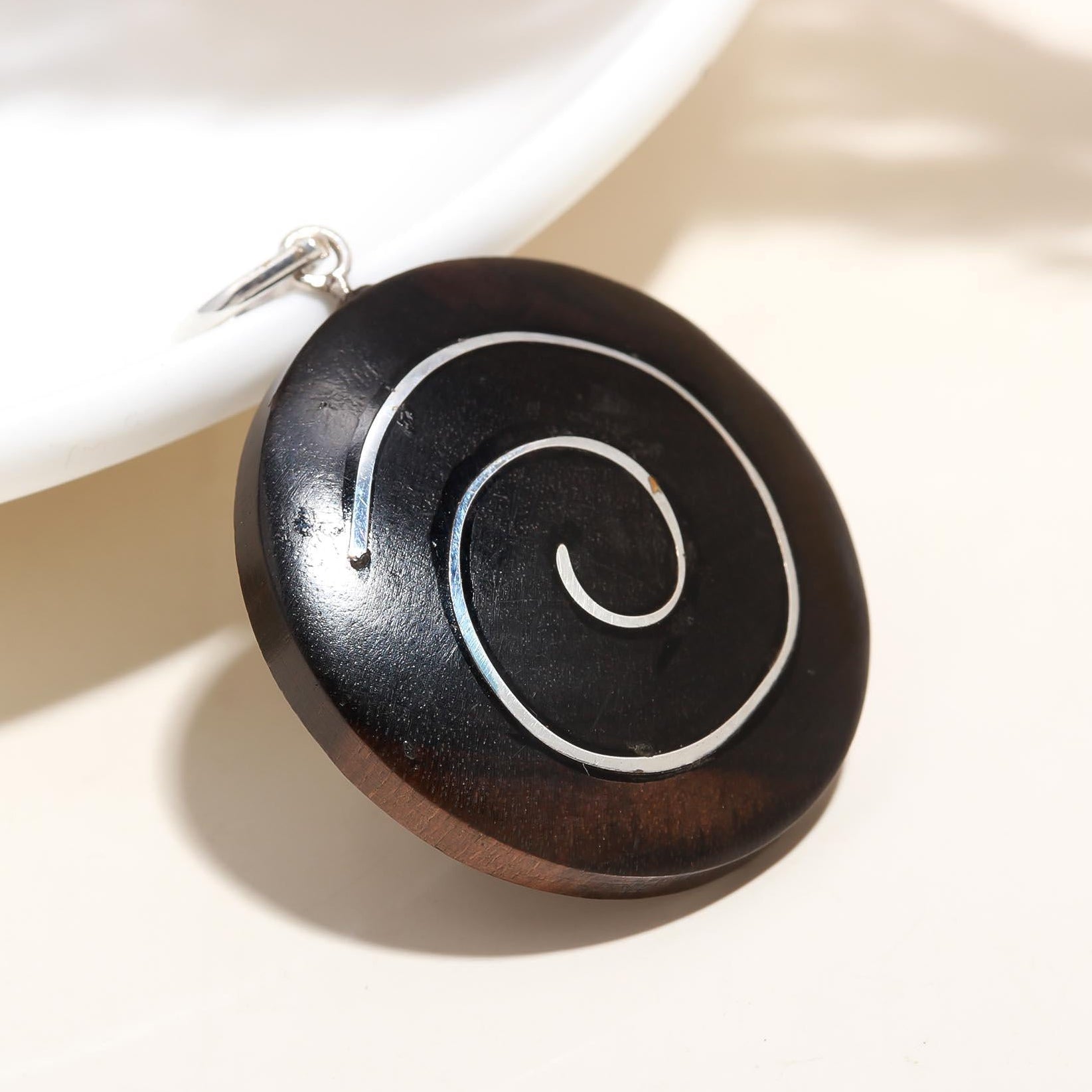 A double-sided ebony wood spiral pendant with a white and blue spiral design on a silver chain, displayed on a light background.