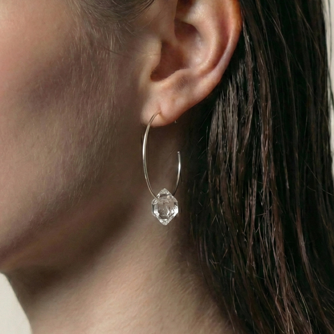 Close-up of a person wearing a silver hoop earring with a clear Herkimer gemstone.