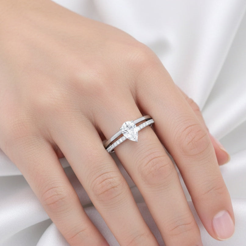 A close-up of a hand wearing a silver ring with a marquise-cut center stone and a matching silver band, set against a soft white satin background.