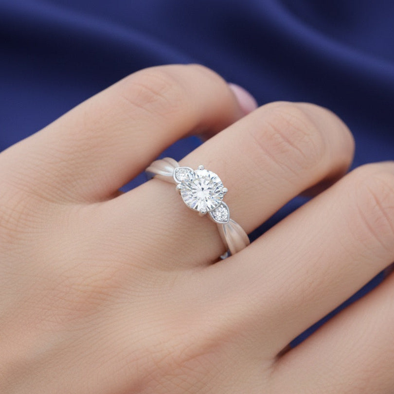 A silver ring with a round-cut center stone and small side stones, worn on a hand against a dark blue satin background.